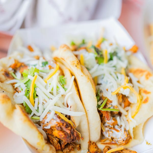 Two chicken flatbread tacos topped with shredded cheese and fresh cilantro in a paper tray.