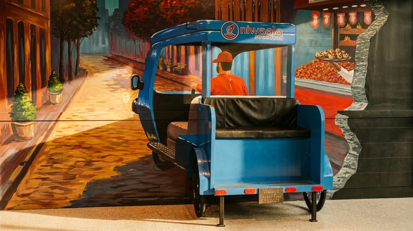 A blue Niwaala Streetfood tuk-tuk is parked against a vibrant mural depicting a street scene and food stall.